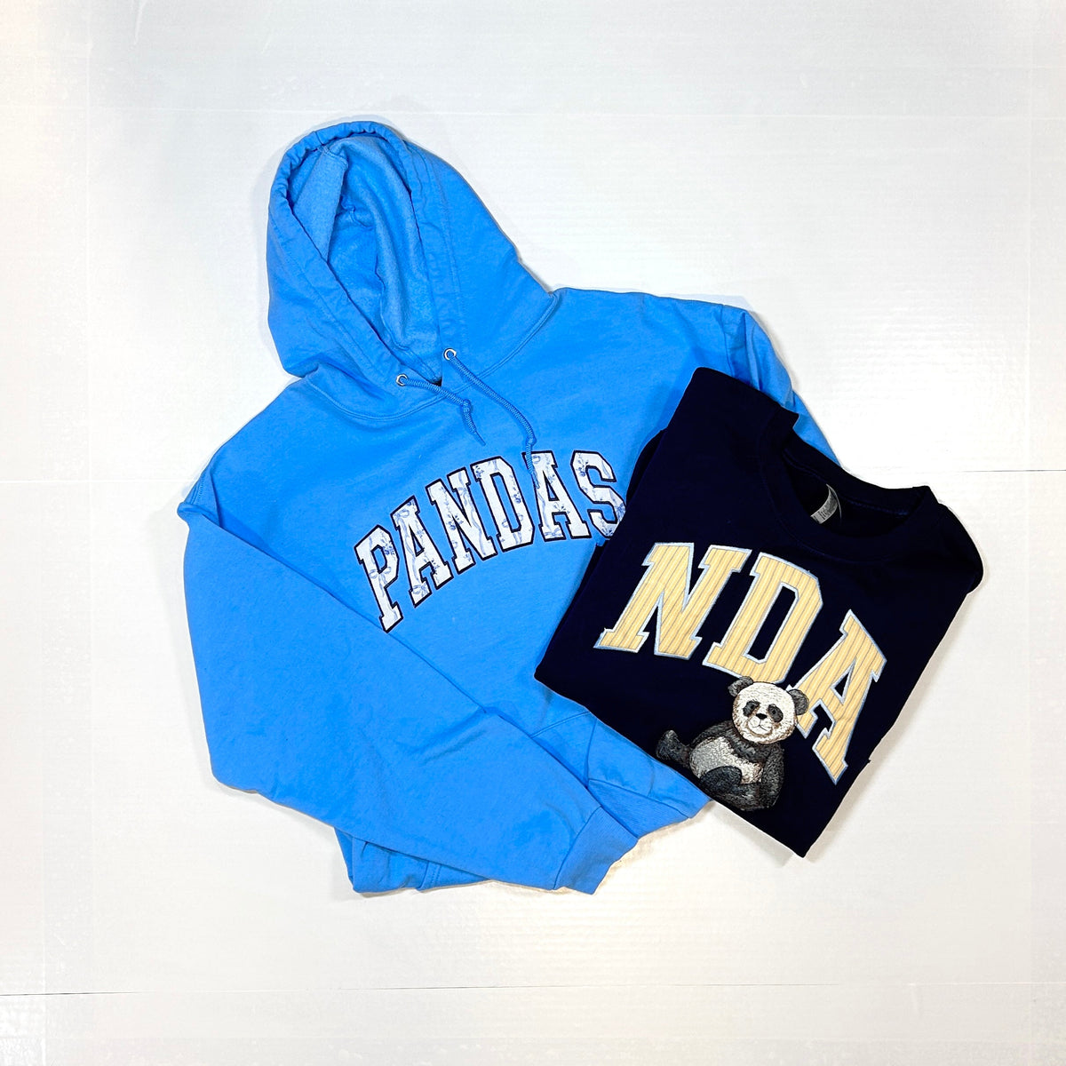 Light blue hoodie with PANDAS fabric appliqué lettering beside a navy NDA crewneck with striped appliqué letters and an embroidered panda.